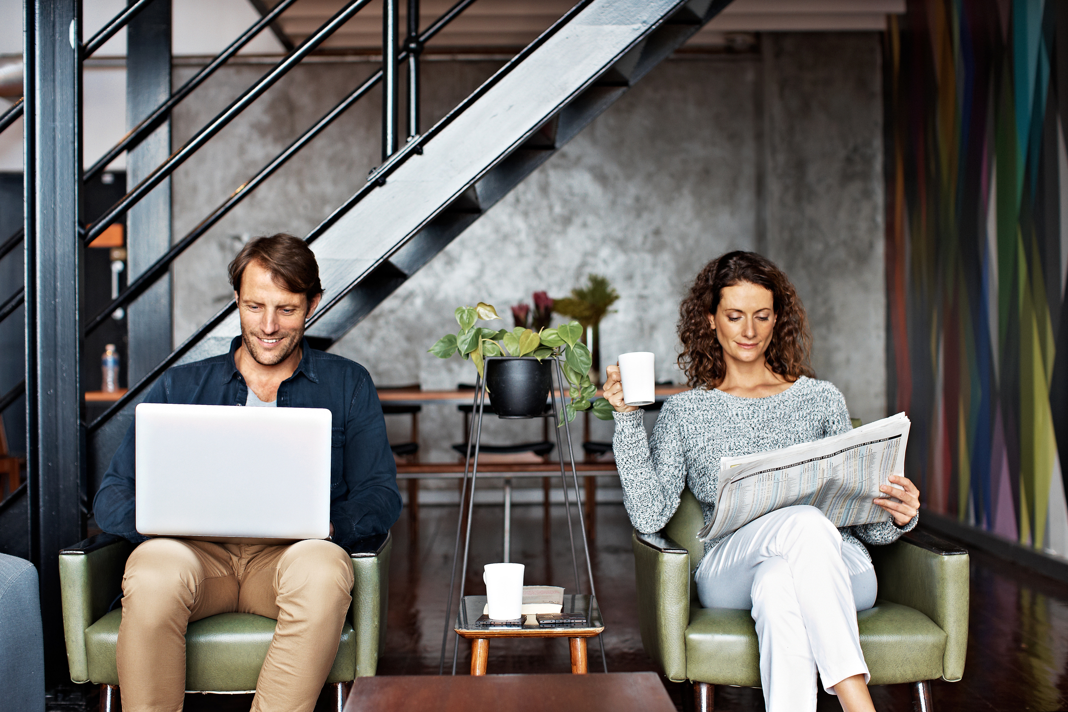 male and female sitting in chairs with one reading on a laptop and the other reading a newspaper