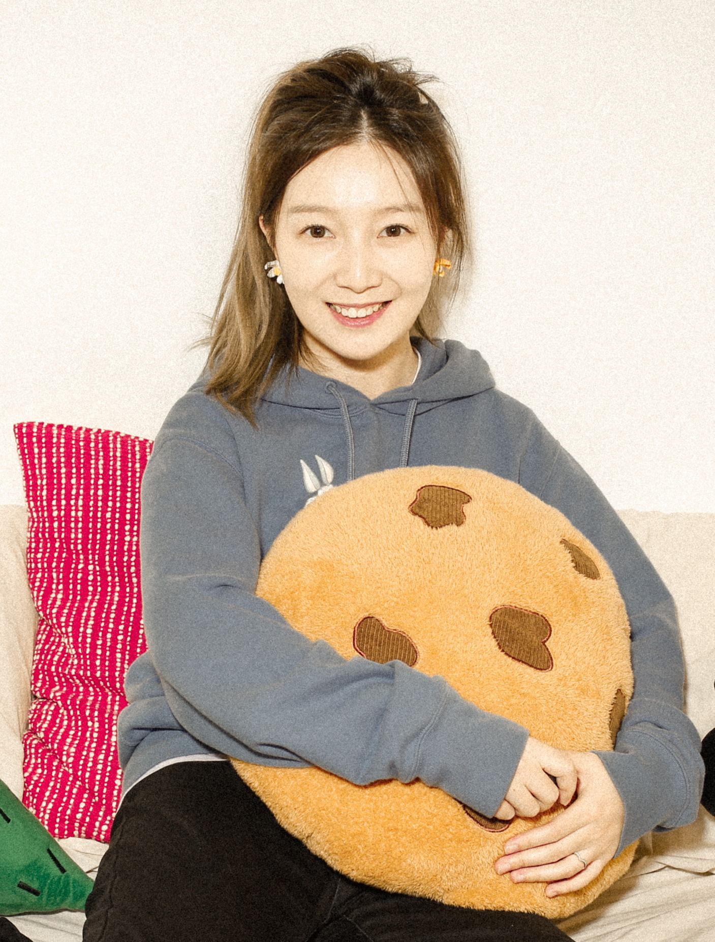 Yie Xie sits on a couch holding what looks like a very snuggly pillow that looks like a giant chocolate chip cookie