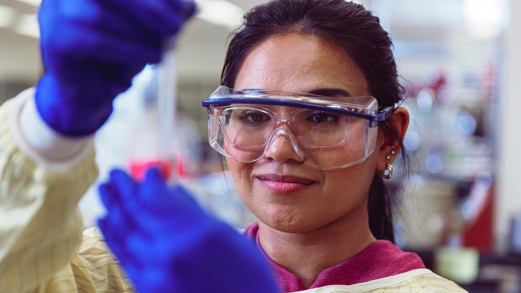 A researcher wearing goggles holds up a test tube in a lab.