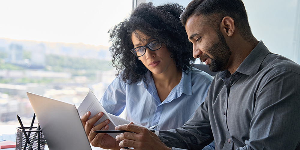 Two people are in front of a laptop pointing at the monitor and looking over documents