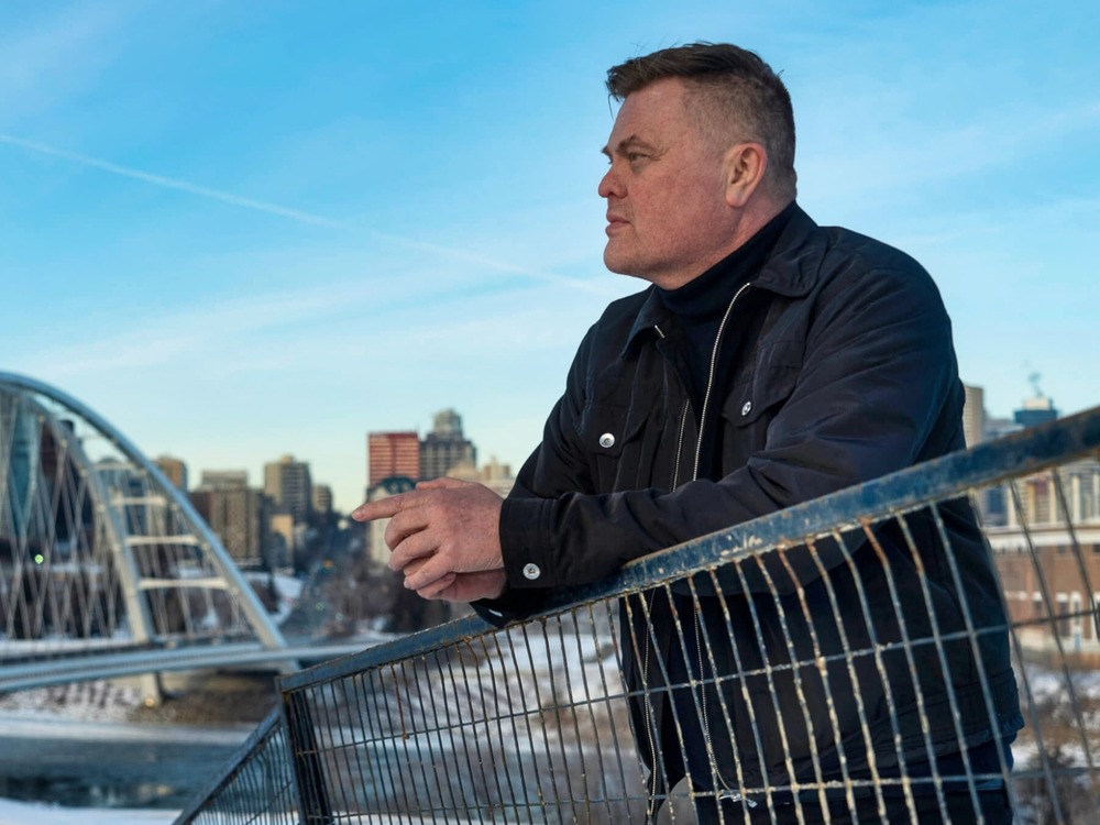 Inuk researcher Wayne Clark leans over a footbridge with the High Level Bridge and downtown Edmonton in the background.