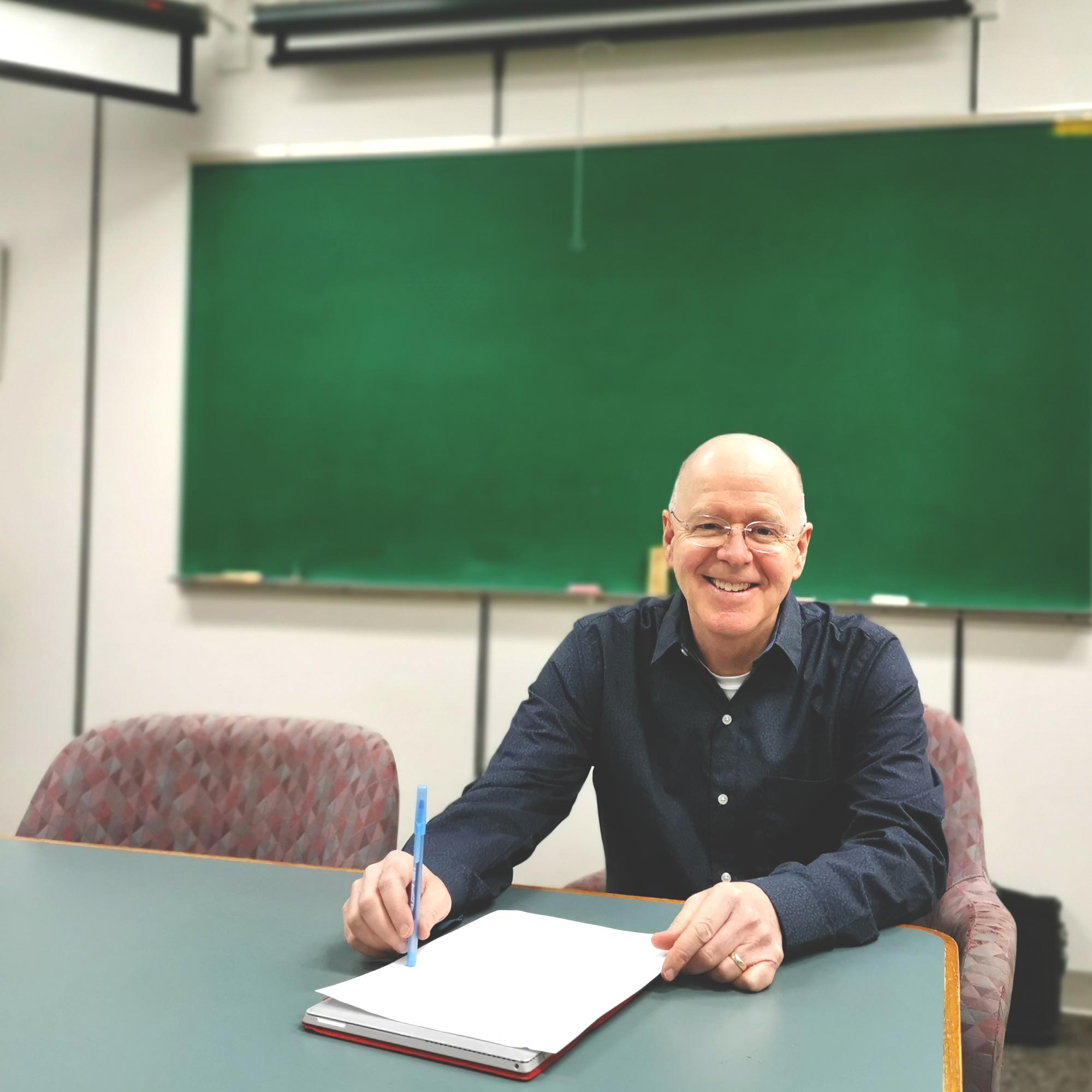 Man smiling behind a table with a computer and paper in front of him